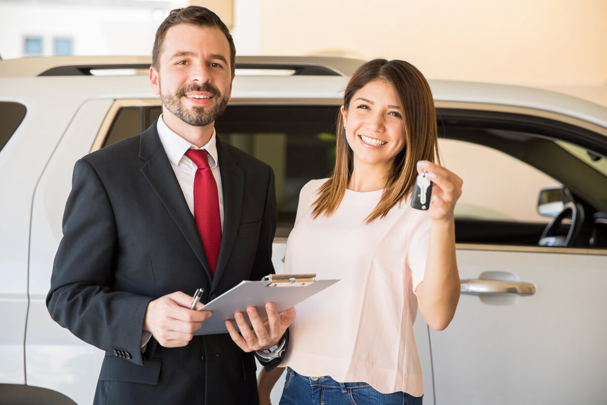 Pretty young woman standing next to a salesman at a dealership and holding the keys to her new car